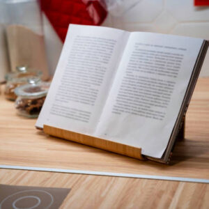 A cookbook standing on top of kitchen table with storage jars by it and a red mitten behind.